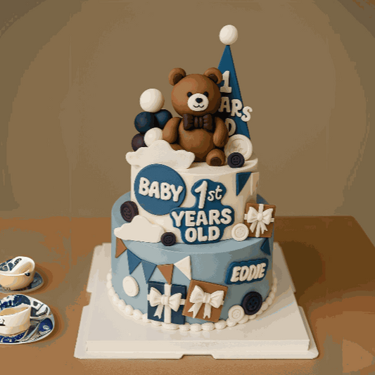 Two-tier Bear Cake in dusty blue and cream with 3D teddy bear topper, fondant gift boxes, custom name plaque, and milestone lettering for baby boy's first birthday celebration