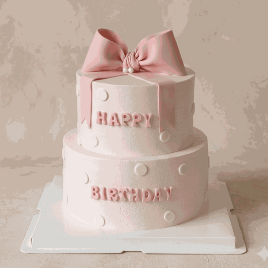 Two-tier pastel pink Bow Cake with white polka dots, 3D fondant bow, and "HAPPY BIRTHDAY" block lettering
