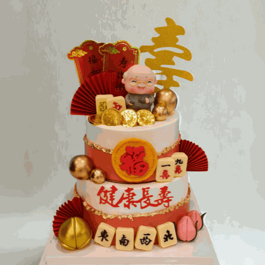 Two-tier Family Cake Longevity Blessing Cake with Shou Star Elder figurine, gold leaf trim, red and ivory design, longevity peaches, mahjong tiles, and Chinese calligraphy banners