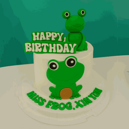Green frog cake with 3D frog topper, happy birthday plaque, and custom lettering on white cream frosting