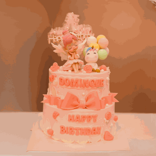 Two-tier unicorn cake with pink fondant bow, sleeping angel figurine, baby unicorn holding pastel balloons, and "Happy Birthday" lettering